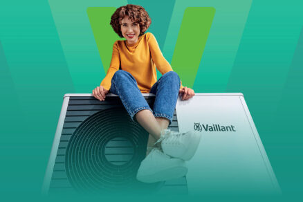 Žena v žltej blúzke a džínsach sedí na tepelnom čerpadle Vaillant aroTHERM plus, na zelenom pozadí s veľkým logom Vaillant.