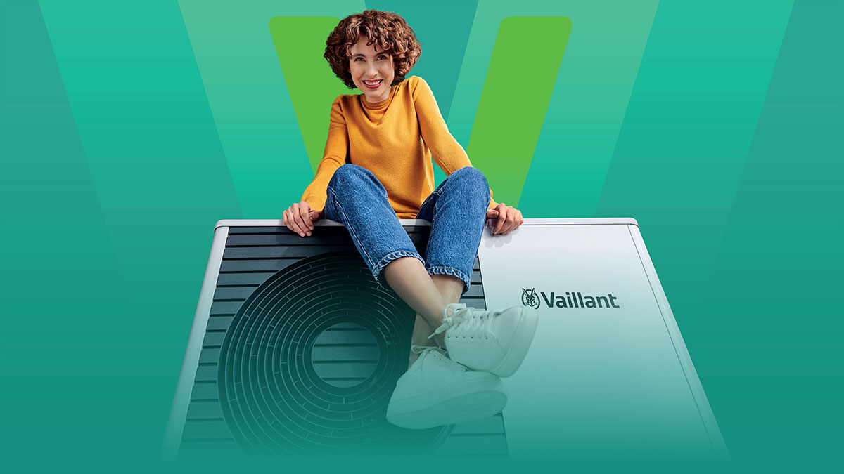  Žena v žltej blúzke a džínsach sedí na tepelnom čerpadle Vaillant aroTHERM plus, na zelenom pozadí s veľkým logom Vaillant.