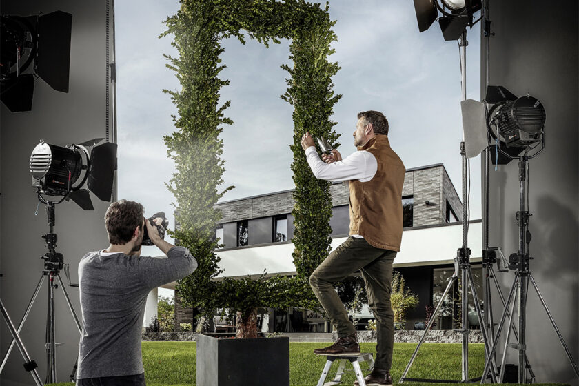 Profesionálny fotograf fotí interiérovú scénu, kde záhradník starostlivo tvaruje živý plot v tvare obrovského obdĺžnika. V pozadí vidno moderný dom a reflektory na osvetlenie scény.