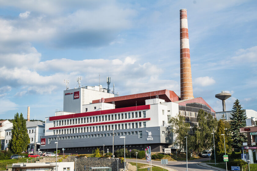 Budova sklárskeho závodu s výrazným červeným komínom a modernou architektúrou, v pozadí jasná obloha s oblakmi.